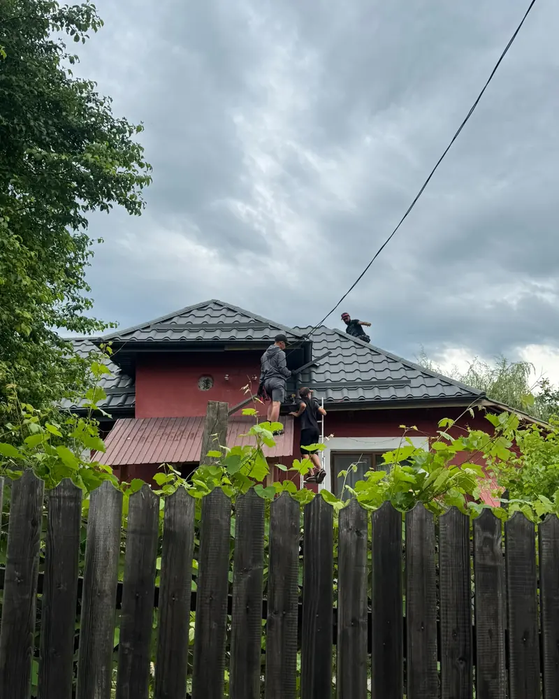 Casă roșie cu acoperiș nou din țiglă metalică și oameni din echipă pe el, în spatele unui gard de lemn